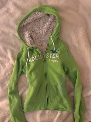Grön Hollister hoodie med teddyfoder - Snygg grön hoodie från Hollister med dragkedja och vit text på bröstet. Insidan av huvan är fodrad med mjukt, vitt teddyfoder. Klassisk logga i blått och vita snören vid huvan. Perfekt för dig som vill ha en mysig och stilren hoodie.