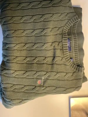 Grön  kabelstickad tröja från GANT - Snygg olivgrön kabelstickad tröja från GANT med rund halsringning och ribbade muddar. Tröjan har GANT:s klassiska logga broderad på bröstet och är tillverkad i mjukt material som passar perfekt till höst och vinter. Den är storlek S men den passar dig som är Xs också. Priset kan diskuteras vid snabb affär