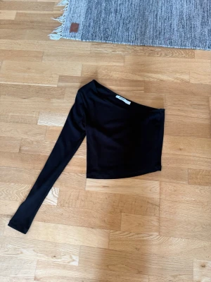 Svart oneshoulder topp från NA-KD - Säljer en svart ribbad oneshoulder topp från NA-KD. Toppen har lång ärm på ena sidan och är croppad i modellen