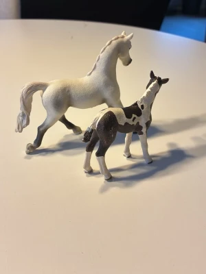 Två hästfigurer i plast - Säljer två detaljerade hästar från Schleich, en helvit och ett brunvitt föl med fläckar.