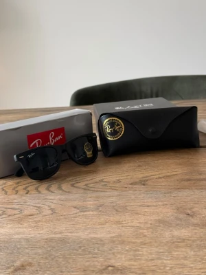 Svarta Ray-Ban Wayfarer solglasögon - Klassiska svarta Ray-Ban Wayfarer solglasögon med tidlös design och mörka glas. Bågarna är i plast och kommer med original svart fodral och Ray-Ban box. Perfekta för dig som vill ha en ikonisk look och skydda ögonen mot solen.