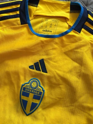 Svensk landslagströja Adidas gul - Gul svensk landslagströja från Adidas med blå detaljer och tre ränder på axlarna. Tröjan har broderat Sverige-emblem på bröstet och blå Adidas-logga. Tillverkad i lätt och ventilerande material, perfekt för fotboll eller träning. Liten i storlek!