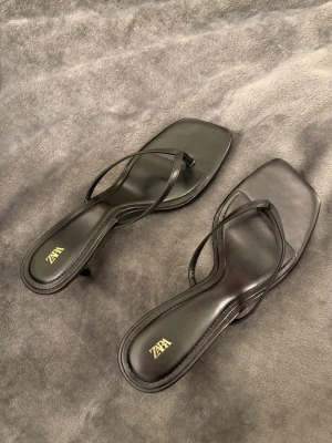 Helt ny Svarta flip flops  med klack från Zara - Helt nya Stilrena svarta flip flops  från Zara med smala remmar och låg klack. Sandalerna har fyrkantig tå och är tillverkade i ett glansigt material som ger en trendig look. Perfekta för dig som gillar minimalistisk och modern stil.