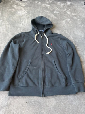 Svart hoodie från Polo Ralph Lauren - Svart hoodie från Polo Ralph Lauren med dragkedja, vit dragsko i huvan och klassisk röd logga broderad på bröstet. Två stora fickor framtill och mjukt material som känns skönt mot huden. Perfekt för en avslappnad och stilren look.