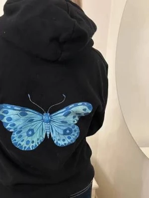 Svart hoodie med blå fjäril - Snygg svart hoodie med stor blå fjäril på ryggen. Hoodien har huva och en avslappnad passform, perfekt för en cool och trendig look. Materialet ser ut att vara mjukt och bekvämt, troligen bomullsblandning. Passar dig som gillar unika tryck och streetwear.