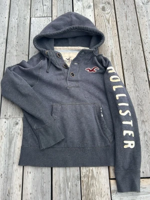 Mörkgrå Hollister hoodie med tryck - Mörkgrå hoodie från Hollister med stor huva, knappar vid halsen och magficka. På vänster ärm står det 'HOLLISTER' i stora bokstäver och på bröstet finns Hollisters logga i rött och vitt. Tillverkad i mjukt sweatshirtmaterial, perfekt för en avslappnad stil.