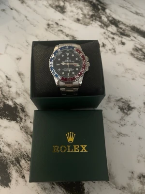 Rolx GMT-Master II Pepsi klocka - Säljer en Rolx GMT-Master II med klassisk Pepsi-randad vridring i blått och rött. Klockan har svart urtavla, silverfärgad länk i rostfritt stål och tydliga indexmarkeringar. Perfekt för dig som gillar ikonisk design och exklusiv känsla.
