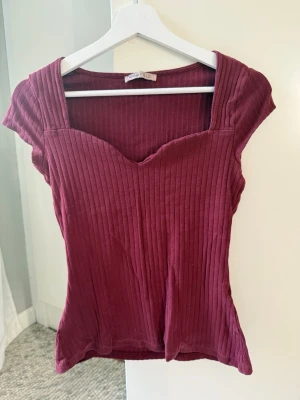 Vinröd ribbad topp med sweetheart-ringning - Säljer en vinröd ribbad topp från Anna Field med snygg sweetheart-ringning och korta ärmar. Toppen har en figurnära passform och är gjord i mjukt, stretchigt material som sitter skönt på kroppen⭐️
