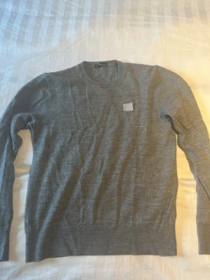 Grå stickad tröja från Acne Studios - Snygg grå stickad tröja från Acne Studios med rund halsringning och diskret logopatch på bröstet. Tröjan har lång ärm och ribbade muddar vid ärmslut och nederkant. Perfekt för lager-på-lager och stilrena outfits.