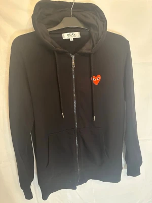 Svart hoodie med rött hjärta - Snygg svart hoodie med dragkedja och huva. På bröstet finns ett litet rött hjärta med ögon, vilket ger en unik och lekfull touch. Hoodien har fickor framtill och justerbara snören i huvan. Perfekt för en chill och avslappnad stil.