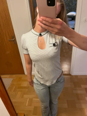 Randig topp med cut-out och knappdetalj - Säljer en vit kortärmad topp som är smalrandig med svarta ränder. Toppen har en unik cut-out. Den har en liten bröstficka och dekorativa svarta knappar vid halsen och fickan. Ärmarna är lite smått puffiga. Nertill går toppen upp lite på sidorna vilket formar kroppen fint. Materialet är mjukt och stretchigt.