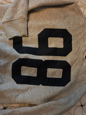 Grå sweatshirt med tryck från Divided - Säljer en grå sweatshirt från Divided med stort svart tryck av siffran 98 på framsidan. Tröjan har rund halsringning och ribbade muddar vid ärmslut och nederkant. Perfekt för en sportig och avslappnad stil.