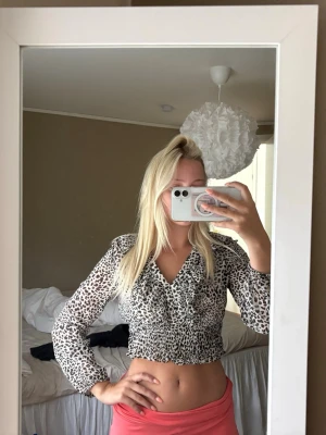 Leopardmönstrad croppad blus - Snygg croppad blus med leopardmönster och v-ringning. Blusen har långa ärmar och smockad midja som ger en tight passform. Perfekt för dig som vill sticka ut med ett trendigt djurmönster.