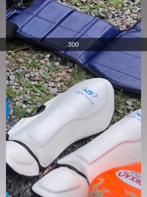 Vita benskydd NH7 för sport - Säljer ett par vita benskydd från NH7, perfekta för sportaktiviteter som fotboll eller innebandy. De har en ergonomisk form och elastiska band för att sitta på plats. Materialet är hårdplast med mjuk insida för extra komfort.