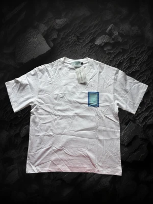Casablanca tröja  - Vit t-shirt med korta ärmar och en blå rektangulär detalj på bröstet. På ryggen finns ett större blått tryck med texten 'CASA BLANCA'. T-shirten har en klassisk passform och är tillverkad i bomull.
