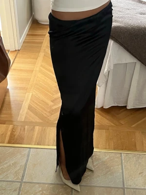 Svart långkjol med slits - Säljer en svart långkjol i ett glansigt material med en slits framtill. Har sytt slitsen själv då den var svår att gå i först, och det kan hända att den fransar lite. Den är svart med en tint av blå.