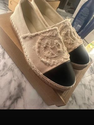 Beige ballerinaskor med svart tå - Säljer ett par trendiga ballerinaskor i beige canvas med fransiga detaljer och svart tåparti. Skorna har en grovsydd sula och ett stort broderat mönster framtill. Perfekta för dig som gillar en avslappnad men ändå stilsäker look.