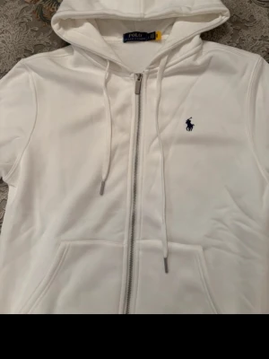 Ralph Lauren Zip Hoodie - Vit hoodie med dragkedja från Polo Ralph Lauren. Klassisk design med blå broderad logga på bröstet. Använd fåtalet gånger snöret är borttaget kommer med köp så man kan sätta dit det. Originalpris 2400 kr Säljs för 499 Funderingar är det bara att skriva!