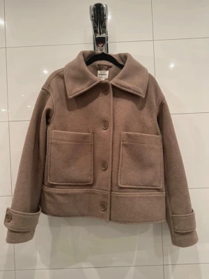 Kort beige/ljusbrun kappa med stora fickor - Snygg beige/brun kappa med rak passform och stora fickor framtill. Kappan har bred krage, knäppning med fyra knappar och dekorativa manschetter vid ärmsluten. Perfekt till hösten🧥