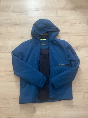 Blå vindjacka från Sail Racing - Snygg blå vindjacka från sail racing med huva och dragkedja framtill. Jackan har en bröstficka med diskret logga och är tillverkad i ett lätt, vindtätt material.
