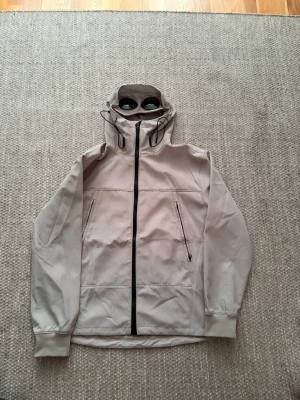 Beige vindjacka från C.P. Company - Snygg beige vindjacka från C.P. Company med ikoniska goggles i huvan. Jackan har hel dragkedja framtill, två snedställda fickor med dragkedja och justerbar huva med snörning. Perfekt för dig som gillar streetwear och unik design.