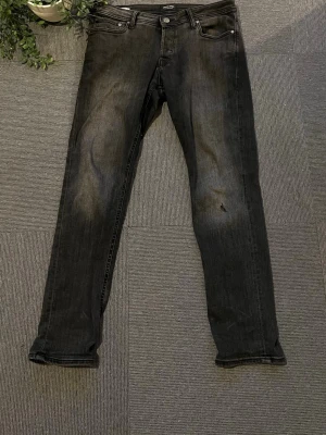 Svart gråa jeans från Jack and Jones  - Snygga svarta slim fit jeans med klassisk femficksdesign och dragkedjegylf. Jeansen har en lätt tvättad look och raka ben. Perfekta för en stilren och avslappnad outfit. Ett litet hål vid vänster knä