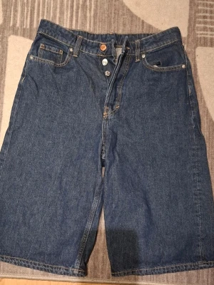 Blå denimshorts med knappar - Säljer ett par klassiska blå denimshorts med knappgylf och fem fickor. Shortsen har en loose passform och är tillverkade i kraftig bomull. Perfekta för en avslappnad stil under varmare dagar.