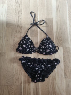 Svart blommig bikini med volang - Söt svart bikini med små blommor i vitt och rosa. Överdelen har halterneck-knyt och volangkant, och bikinitrosorna har matchande volangdetaljer med scrunch I bak. Överdelen är I M och underdelen är I S. Från cubus. Nypris för båda är ca 330. Man kan ta ur inläggen ur den! Några sömmar i underdelen har gått upp men det syns inte om man har på sig den, skriv om ni vill ha bilder!🫶🏻