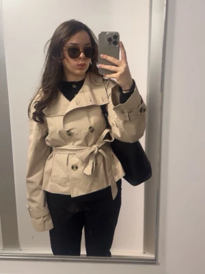 Beige trenchcoat från Bershka - Stilren beige trenchcoat från Bershka med knytskärp i midjan och bred krage. Jackan har dubbelknäppning och är tillverkad i ett lätt, slätt material som passar perfekt till vår och höst. Klassisk och tidlös design som funkar till många olika looks.