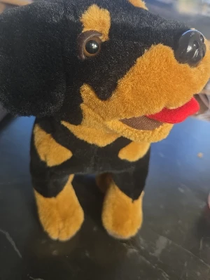 Mjukisdjur hund svart och brun - Söt mjukishund i svart och brunt plyschmaterial med bruna detaljer och röd tunga. Hunden har stora ögon, mjuk nos och står på fyra ben. Perfekt som gosedjur eller dekoration. Ingen synlig märkning.