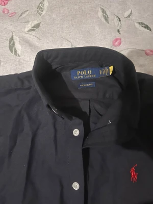 Mörkblå skjorta från Polo Ralph Lauren - Klassisk mörkblå skjorta från Polo Ralph Lauren med vit knappstängning och röd broderad logga på bröstet. Skjortan har lång ärm och en slim fit passform som ger en clean och stilren look. Perfekt till jeans eller chinos.