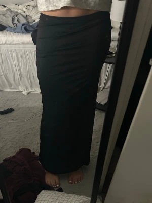 Svart långkjol med slits - Säljer en svart långkjol med slits bak. Kjolen har en tight passform och är gjord i ett mjukt, stretchigt material som sitter snyggt längs kroppen. Perfekt att styla med en croppad tröja eller en enkel topp.
