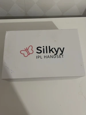 Silkyy IPL Handset hårborttagning - Silkyy IPL Handset för hårborttagning hemma. Setet innehåller en svart IPL-enhet med gulddetalj, skyddsglasögon och vit strömkabel. Perfekt för smidig och enkel hårborttagning med modern teknik.