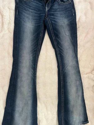 Blå bootcut jeans med broderade fickor - Säljer ett par blå bootcut jeans med snygga slitningar och kontrastsömmar. Bakfickorna har broderade detaljer och fransade kanter som ger en cool look. Jeansen har normal midja och är tillverkade i klassisk denim.