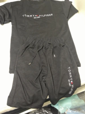 Svarta shorts från Tommy Hilfiger - Säljer ett par svarta shorts från Tommy Hilfiger med snörning i midjan och logga tryckt på ena benet. Shortsen har en avslappnad passform och är perfekta för sommaren. Materialet känns mjukt och bekvämt, perfekt för chill dagar.