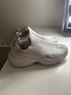 Vita slip-on sneakers från Aimn - Säljer ett par vita slip-on sneakers från Aimn med chunky sula och stickad ovandel i mesh. Skorna har elastisk resår runt öppningen och en svart logga på hälen. Perfekta för en sportig och trendig look. Har använt de inne på gymmet endast. 