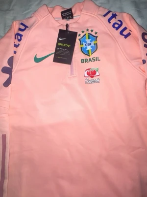 Nike Brasil träningströja rosa - Säljer en rosa träningströja från Nike med Brasilien CBF-logga, grönt Nike-märke och Guaraná Antarctica-tryck. Tröjan har halv dragkedja, tryck på ärmarna och är tillverkad i ett lätt, andningsbart material. Perfekt för fotbollsträning eller sportiga dagar.