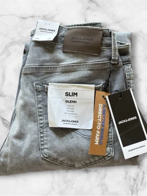 Slim fit grå jeans från Jack & Jones - Slim fit jeans i ljusgrå tvätt från Jack & Jones, modell GLENN. Byxorna har låg midja, smala ben och klassisk femficksdesign. Tillverkade i stretchigt material för extra komfort och snygg passform. Perfekta för en trendig och avslappnad stil. Oanvända