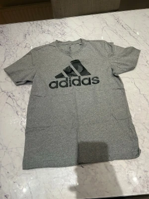 Grå Adidas t-shirt med logga - Snygg grå t-shirt från Adidas med stor svart logga på bröstet. Klassisk passform och rund halsringning. Perfekt till vardags eller träning. Tillverkad i mjuk bomull för skön känsla.