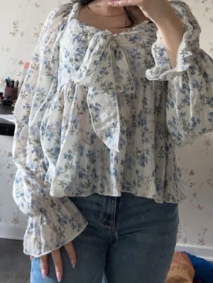 Blommig volangblus med rosett - Romantisk blus med volanger och stor rosett framtill. Blusen har ett blommigt mönster i blått och grönt på vit botten, samt långa, vida ärmar med resår vid ärmslut. Tillverkad i ett lätt, transparent tyg som ger en luftig känsla.