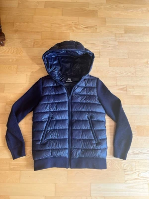 Mörkblå pufferjacka/cardigan med huva från maggiore  - Snygg mörkblå pufferjacka/cardigan med stickade ärmar och huva. Jackan har quiltad front och rygg i glansigt material, dragkedja framtill och två fickor med dragkedja. Perfekt för kalla dagar och ger en sportig look.                  JAG KÖPTE DEN FÖR 1799kr. MITT PRIS 1399        Har använts upp mot fem gånger 