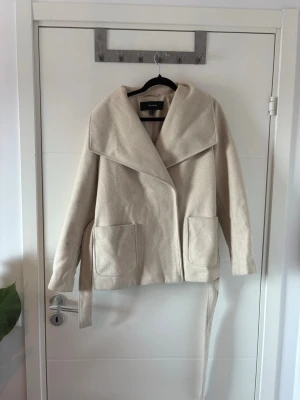 Beige kappa från Vero Moda - Vero Moda jacka, stl M  Kort jacka i ljusbeige med stor krage, uttagbart knytband och två knappar framtill. Fint använd, men har tecken på användning i form av noppor på tyget. I övrigt hel och funktionell.                                                  Har bundle-rabatt så kolla gärna in min sida, buda gärna🧡