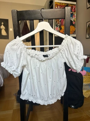 Vit offshoulder blus från Gina Tricot - Söt vit offshoulder blus från Gina Tricot med broderade hål och volangdetaljer. Blusen har puffiga korta ärmar och resår vid axlar och midja för en bekväm passform. Perfekt för sommarens alla tillfällen!