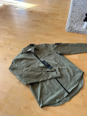 Olivgrön overshirt från Stone Island - Snygg olivgrön overshirt från Stone Island med dragkedja framtill, bröstficka med lock och klassisk krage. Tillverkad i ett slitstarkt bomullsmaterial och har den ikoniska loggan på ärmen. Perfekt för lager på lager och streetwear-stil.