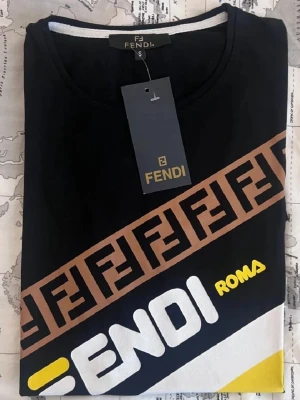 Svart Fendi t-shirt med logga - Snygg svart t-shirt från Fendi med diagonal brun loggoremsa, stora vita och gula tryck med texten 'Fendi Roma' och färgblock i vitt och gult. Rund halsringning och klassisk passform. Perfekt för dig som gillar streetwear och exklusiva märken.