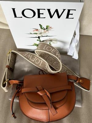 Brun axelväska i läder från Loewe - Snygg brun axelväska från Loewe i slätt läder med dekorativ knut framtill och bred beige axelrem med logotyp. Väskan har flera innerfack och magnetlås. Perfekt för dig som vill ha en trendig och exklusiv accessoar. Kommer i original låda med dustybag och påse . 