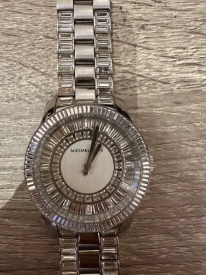 Silverfärgad klocka Michael Kors - Säljer en snygg silverfärgad klocka från Michael Kors med stenar  runt urtavlan och på armbandet. Klockan har en klassisk rund form och länkarmband i metall som ger en lyxig känsla. Perfekt accessoar för att lyfta din stil. Klockan har dock gått sönder men går att laga (se sista bilderna) och batteriet behöver bytas ut!