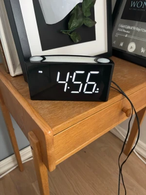 Digital väckarklocka med LED-display - Snygg digital väckarklocka med stor vit LED-display och svart rektangulärt hölje. Klockan har två stora knappar på ovansidan och en inbyggd lampa som kan lysa i olika färger, t.ex. lila. Perfekt för nattduksbordet och enkel att använda med sladdanslutning. Finns även en vibrations funktion med den svarta bollen. 
