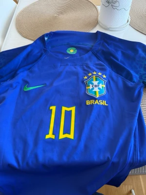 Blå Brasilien Neymar Jr. matchtröja - Säljer en blå Brasilien fotbollströja från Nike med Neymar Jr. och nummer 10 på ryggen. Tröjan har gula detaljer, på bröstet och grönt Nike-märke. Ärmarna har ett mönster i blått och grönt. Perfekt för dig som älskar fotboll!