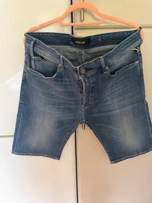 Blå jeansshorts från Replay - Säljer ett par klassiska blå jeansshorts från Replay med fem fickor och dragkedja. Shortsen har en normal passform och är tillverkade i mjuk bomull med snygga slitningar och tvättade detaljer.Längd 55 cm.  Bredd midjan 96 cm, bredd längst nere 48 cm.  Nya och oanvända